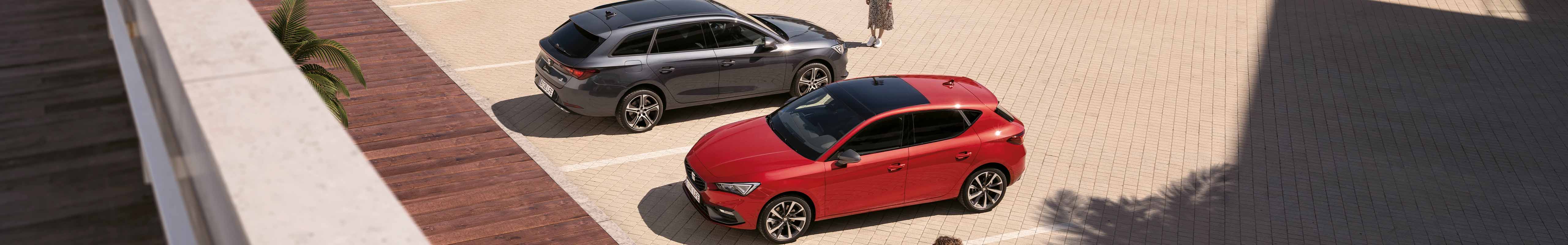 Vue aérienne de deux SEAT Leon, l’une en Magnetic Tech Grey et l’autre en Desire Red, garées devant une femme.