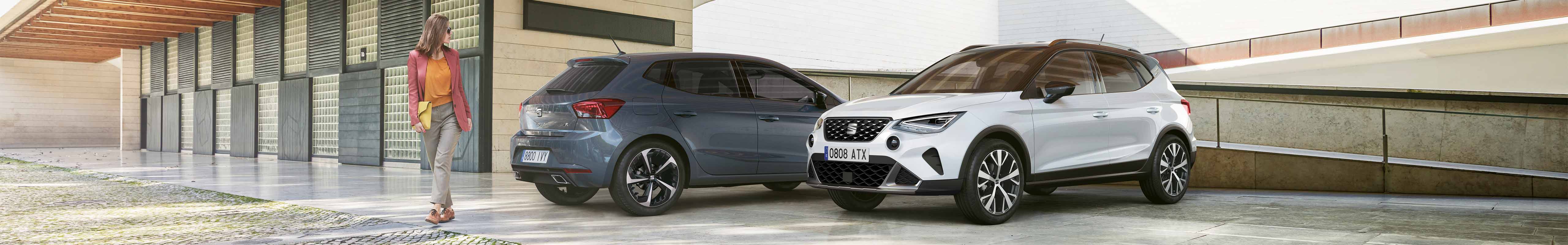 Une femme se promenant entre une SEAT Ibiza en Magnetic Tech Blue et une SEAT Arona en Nevada White et Midnight Black, après avoir effectué les mises à jour OTA